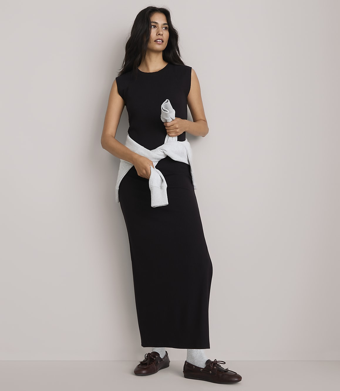 A woman in a black maxi dress and light blue sweater tied around her waist stands against a plain, light gray background.