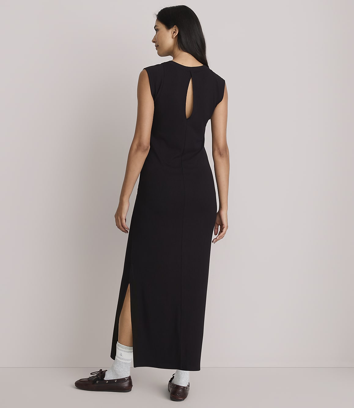 A woman in a black maxi dress with a cutout back and side slits stands with her back to the camera, wearing white socks and brown loafers against a plain, light-colored background.
