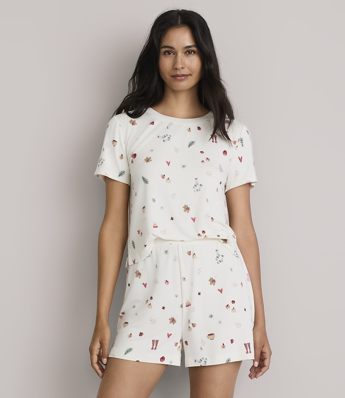 A woman in a cream-colored pajama set with a scattered pattern of small festive icons stands against a plain, light-colored background.
