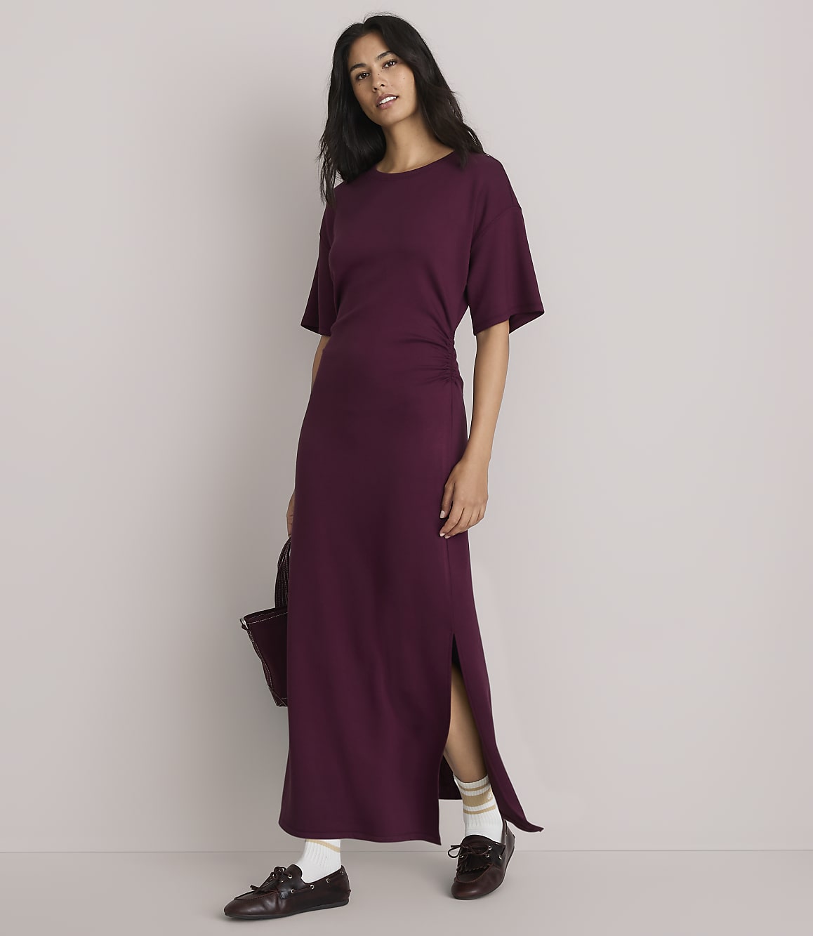 A woman in a plum-colored maxi dress with a side slit stands against a plain, light gray background, holding a dark bag.