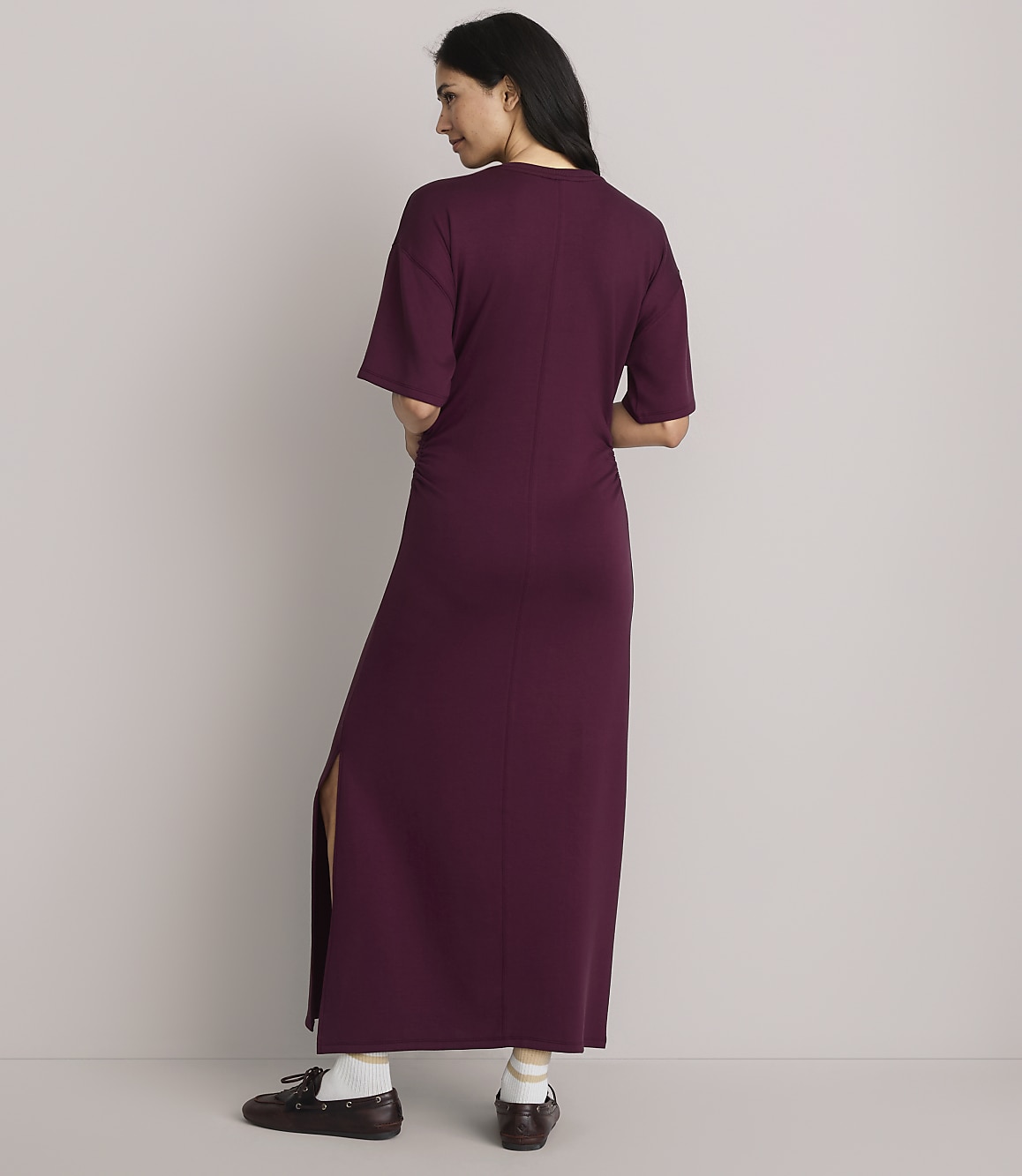 A woman in a burgundy maxi dress with a side slit stands against a plain gray background.