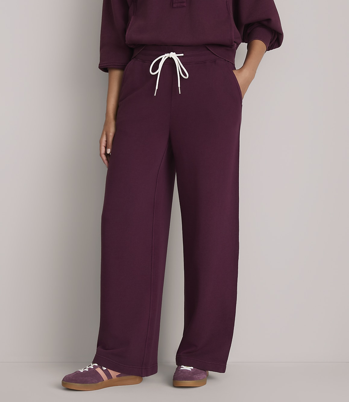 A person wearing burgundy sweatpants with a white drawstring stands against a plain gray background, with their feet in burgundy sneakers.