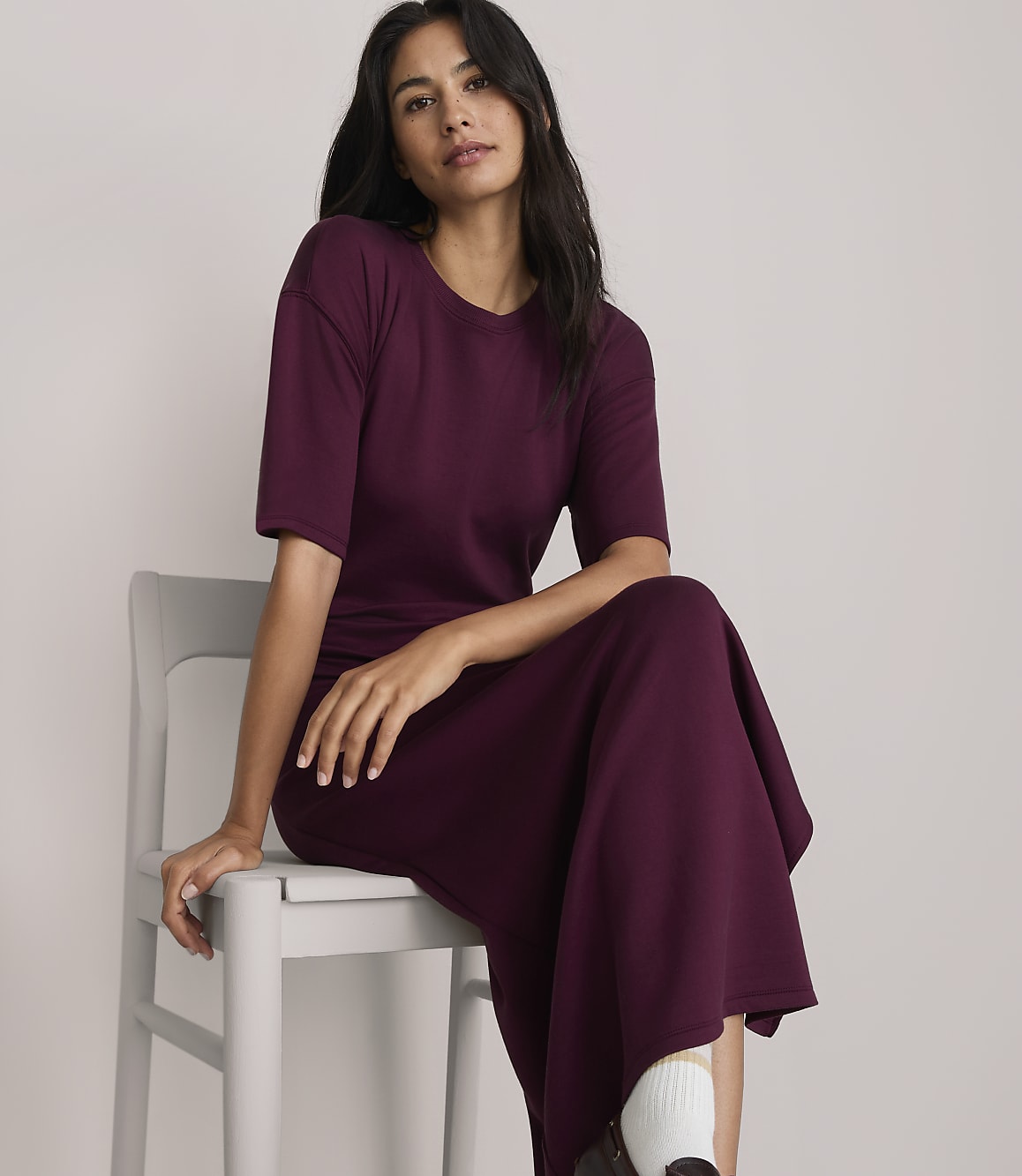 A woman in a burgundy dress sits on a gray chair against a plain white background.