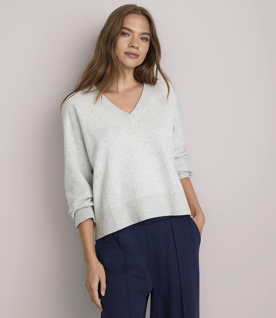 A woman in a light gray v-neck sweater and navy blue pants stands against a plain white background.