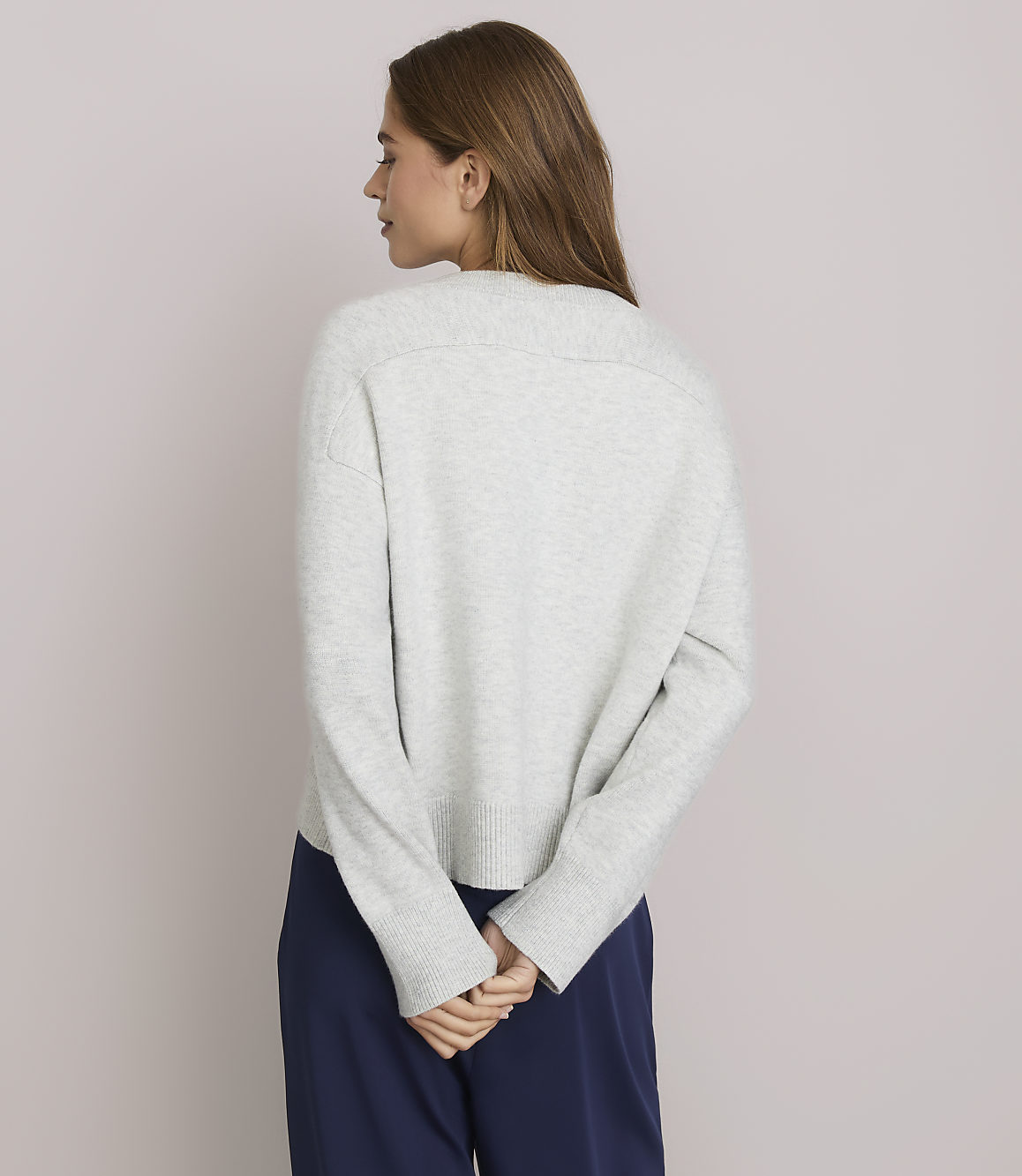 A woman in a light gray sweater and navy pants stands with her back to the camera against a plain white background.