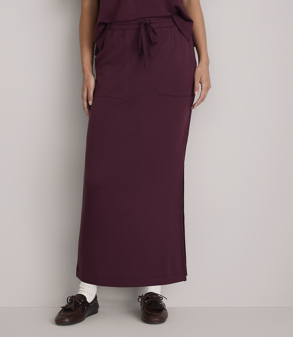 A person wearing a burgundy maxi skirt and brown loafers stands against a plain white background.