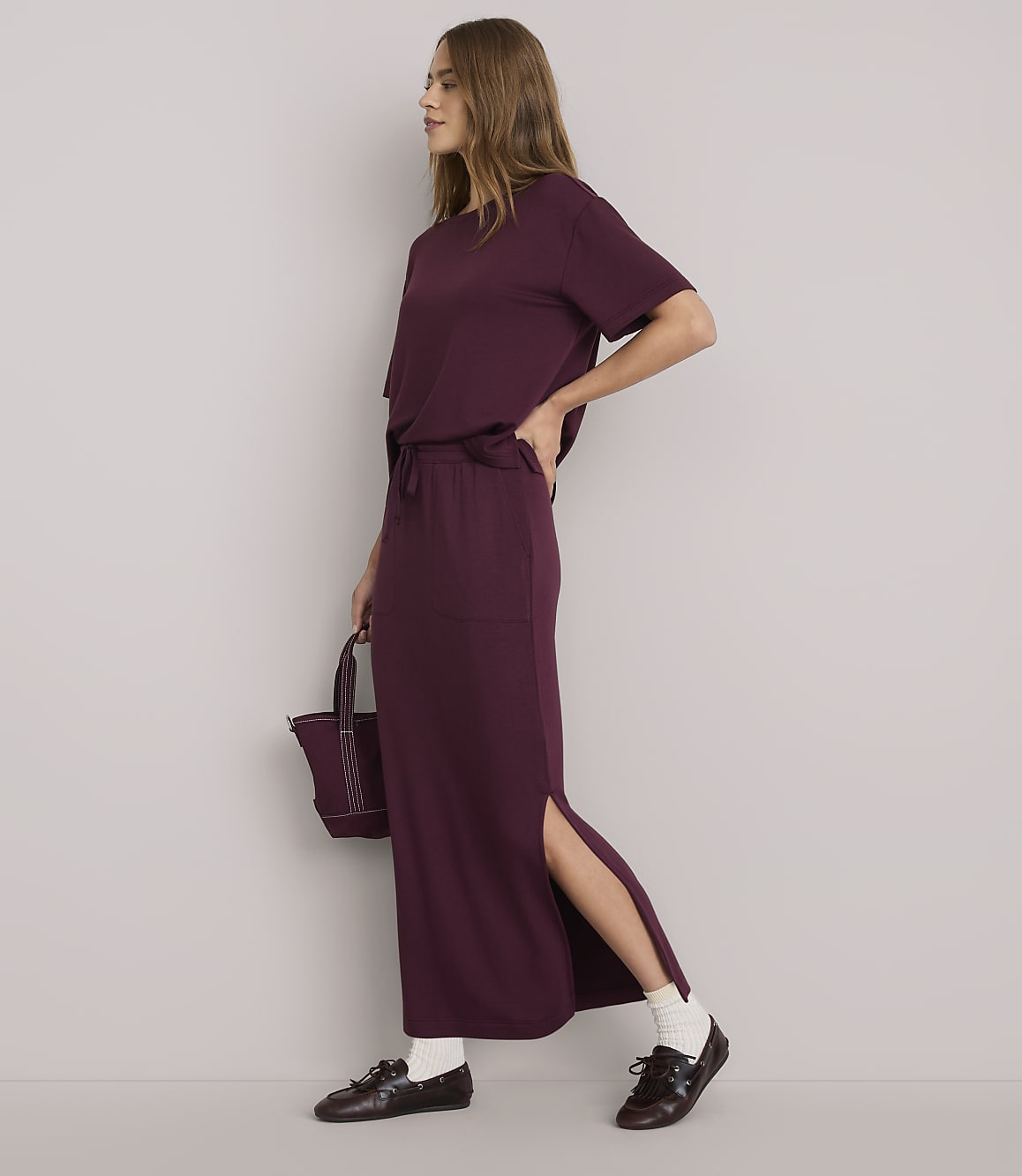 A woman in a burgundy t-shirt and matching maxi skirt with a side slit stands against a plain white background, holding a small burgundy bag.
