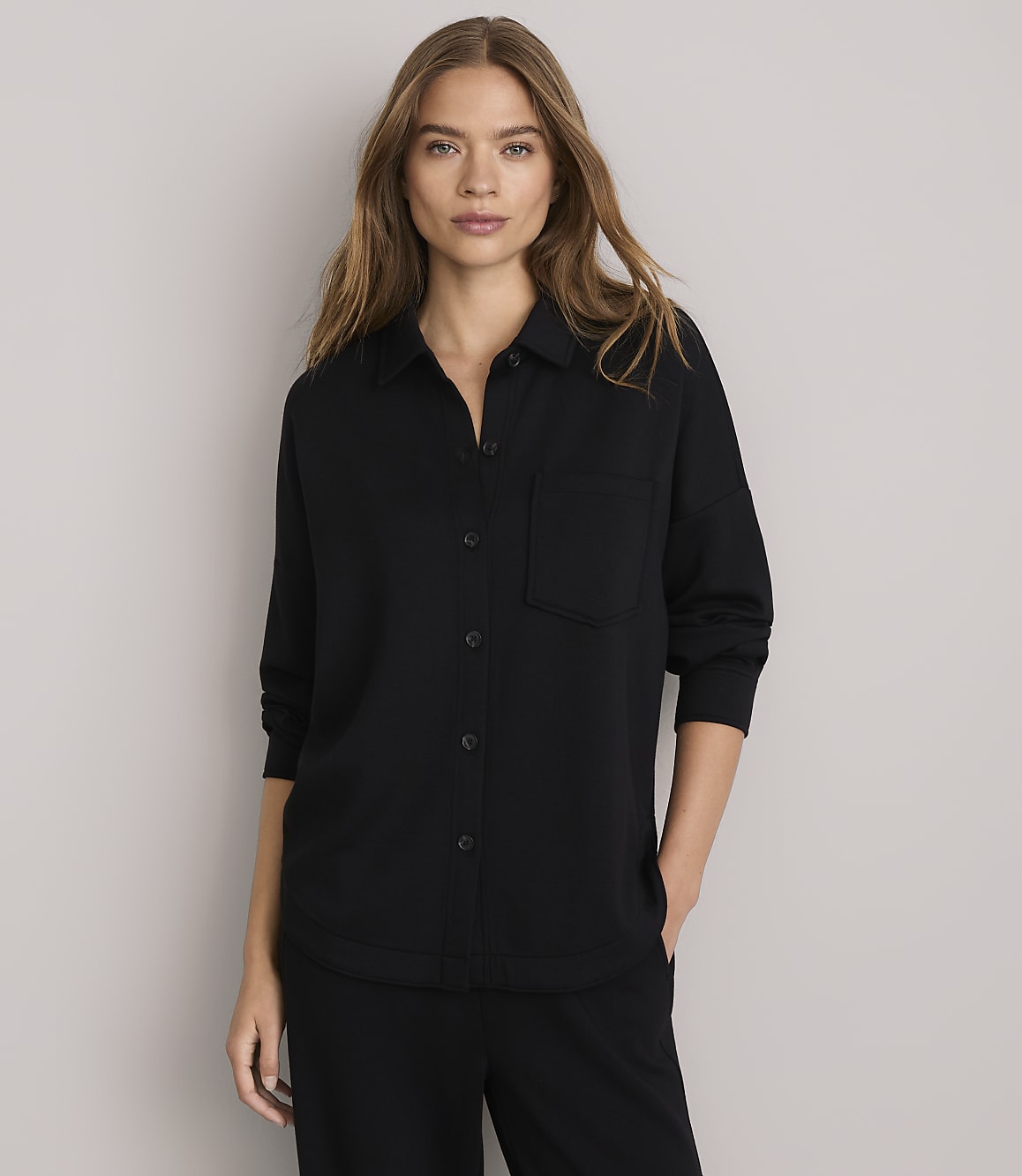A woman in a black collared shirt and pants stands against a plain gray background.