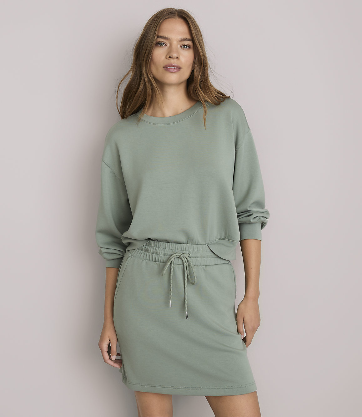 A woman in a green sweatshirt and skirt stands against a plain white background.