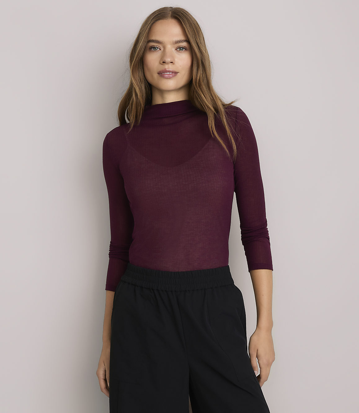 A woman in a burgundy turtleneck and black pants stands against a plain white background.