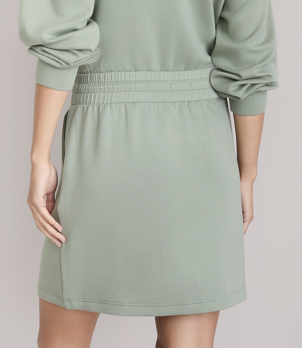 A person in a light green skirt and sweatshirt stands against a plain white background.