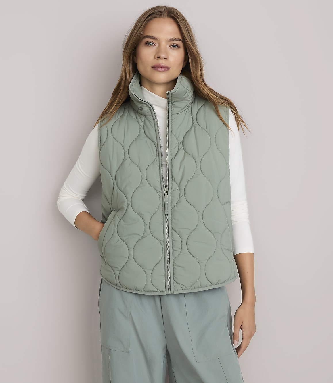 A woman in a light green quilted vest and white turtleneck stands against a plain white background.