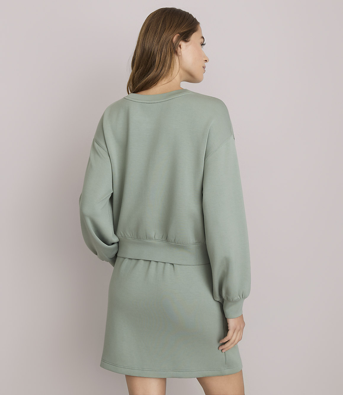 A woman in a sage green sweatshirt and skirt stands against a plain white background.