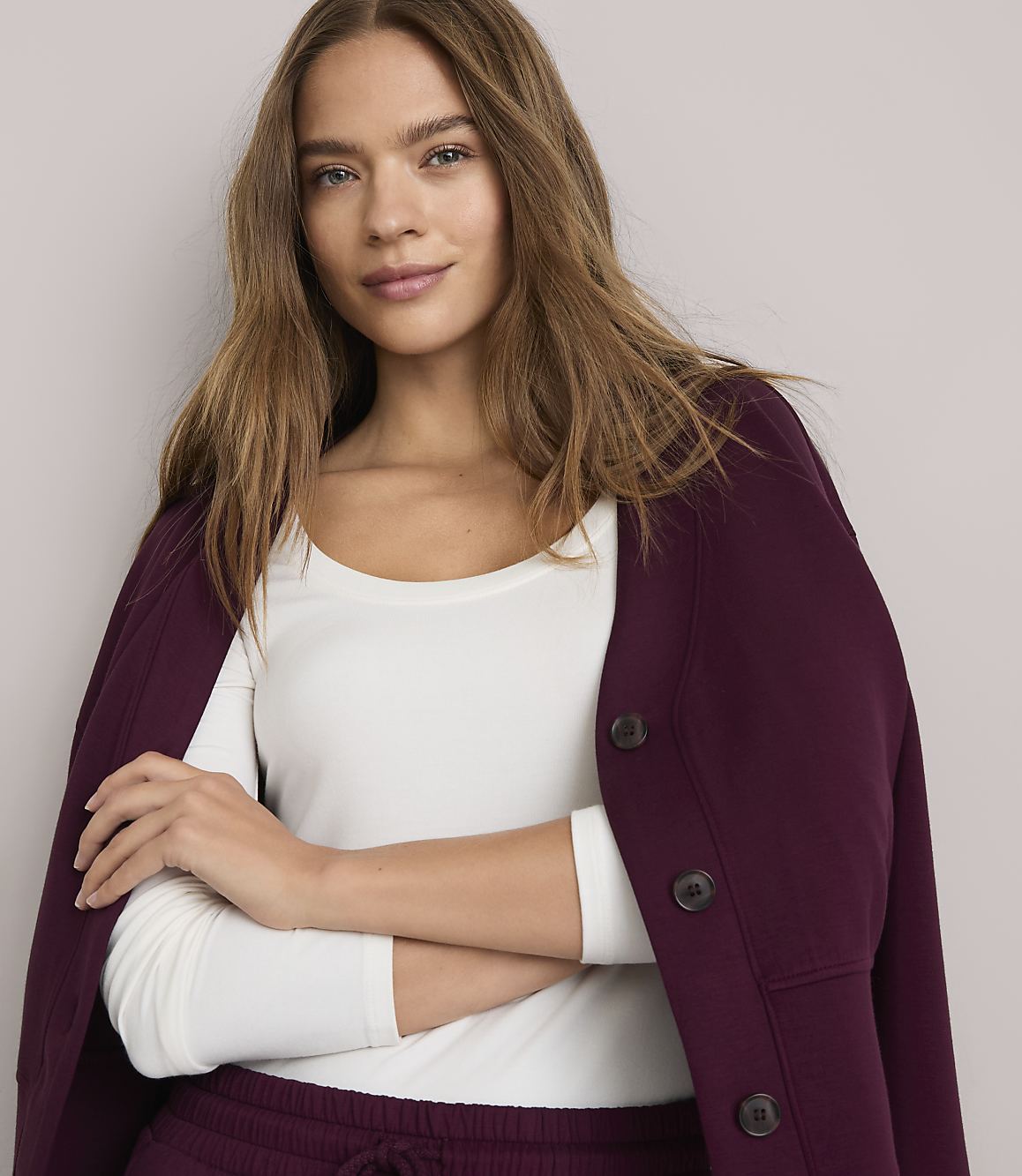 A woman in a white long-sleeved shirt and a burgundy cardigan stands against a plain gray background.