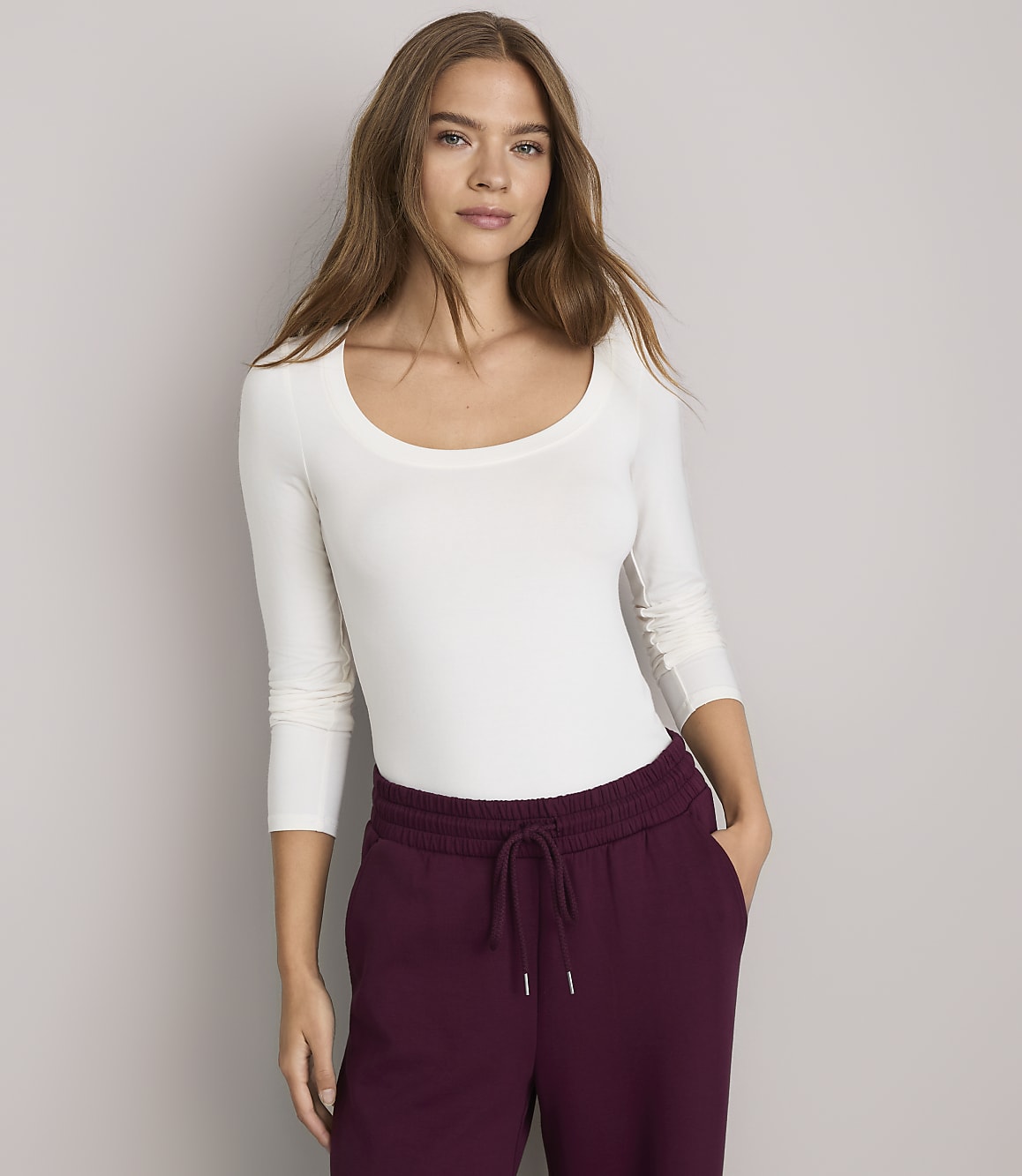 A woman in a white long-sleeved shirt and burgundy sweatpants stands against a plain gray background.