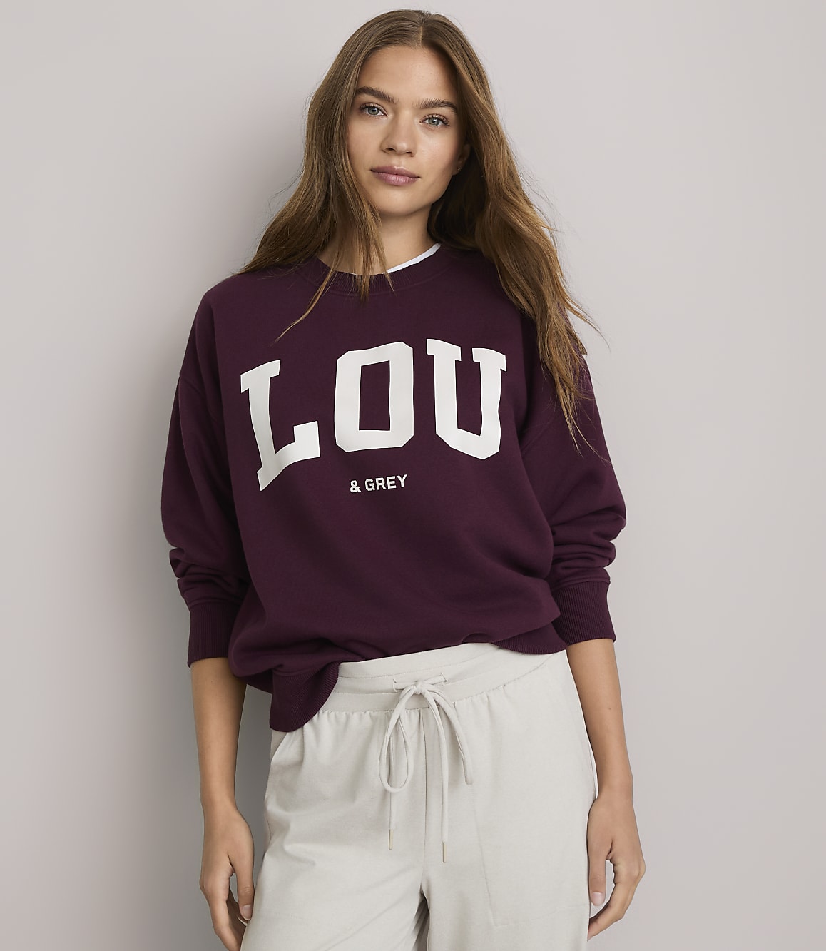 A young woman in a maroon sweatshirt with "LOU & GREY" printed on it stands against a plain white background.