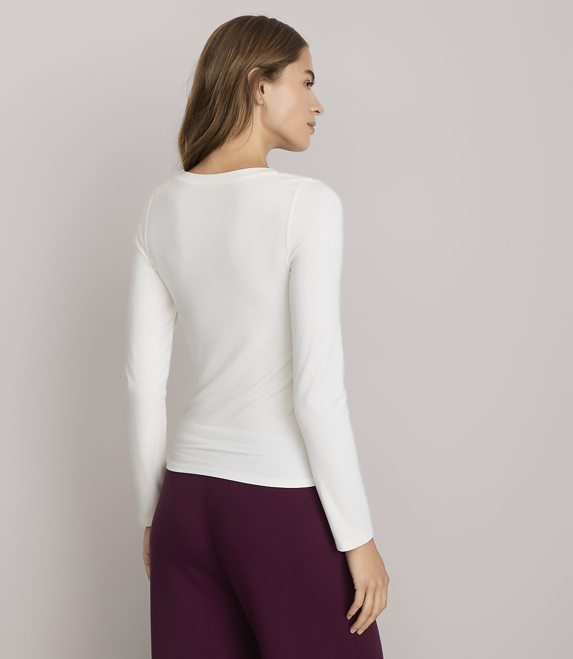 A woman in a white long-sleeved shirt and burgundy pants stands against a plain gray background.