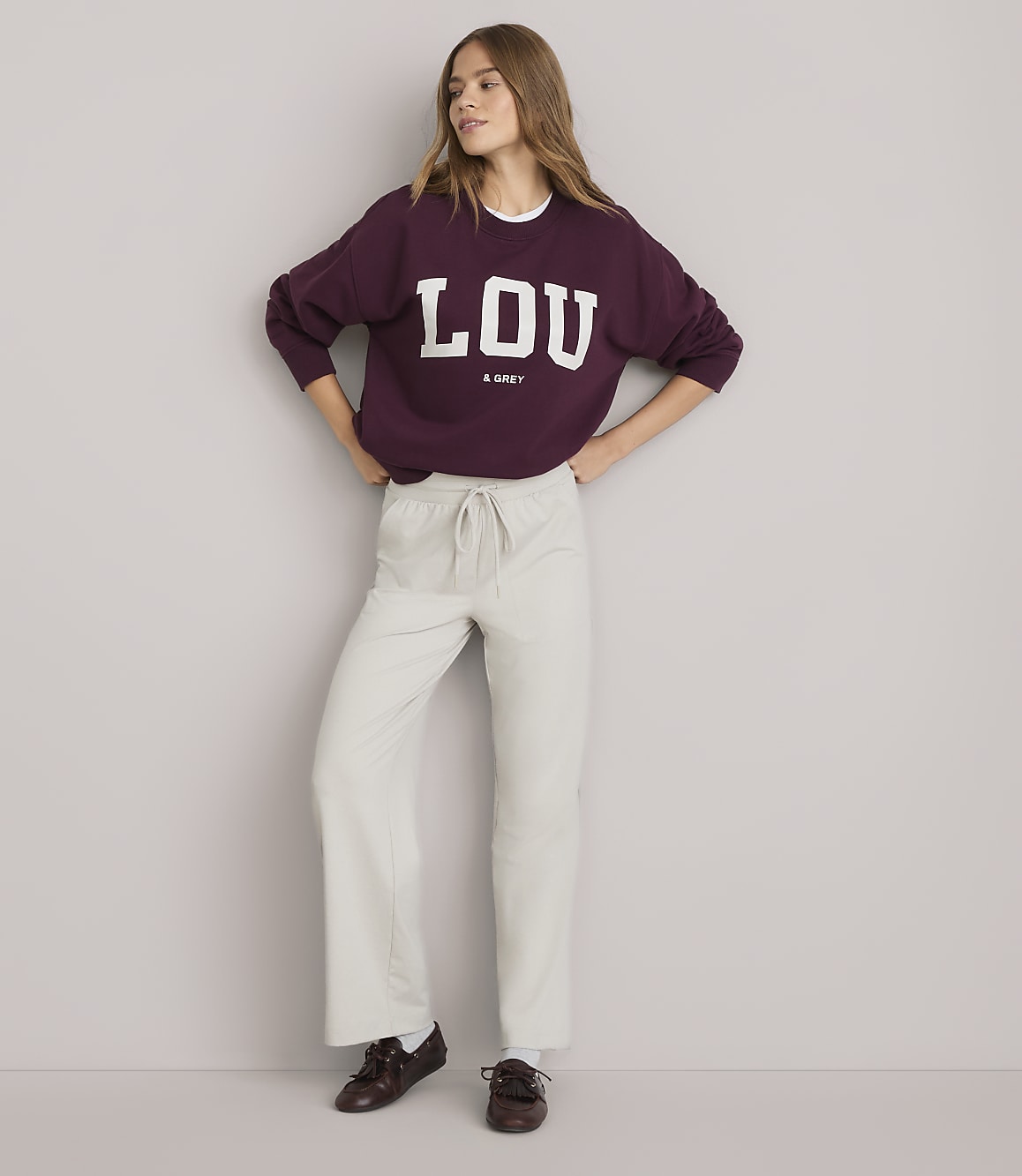 A young woman in a maroon sweatshirt and light-colored pants stands against a plain white background.