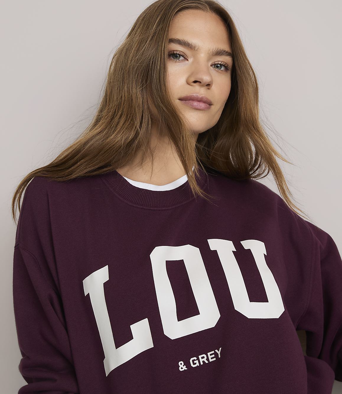 A young woman in a maroon sweatshirt with "LOU & GREY" printed on it is shown against a plain, light-colored background.