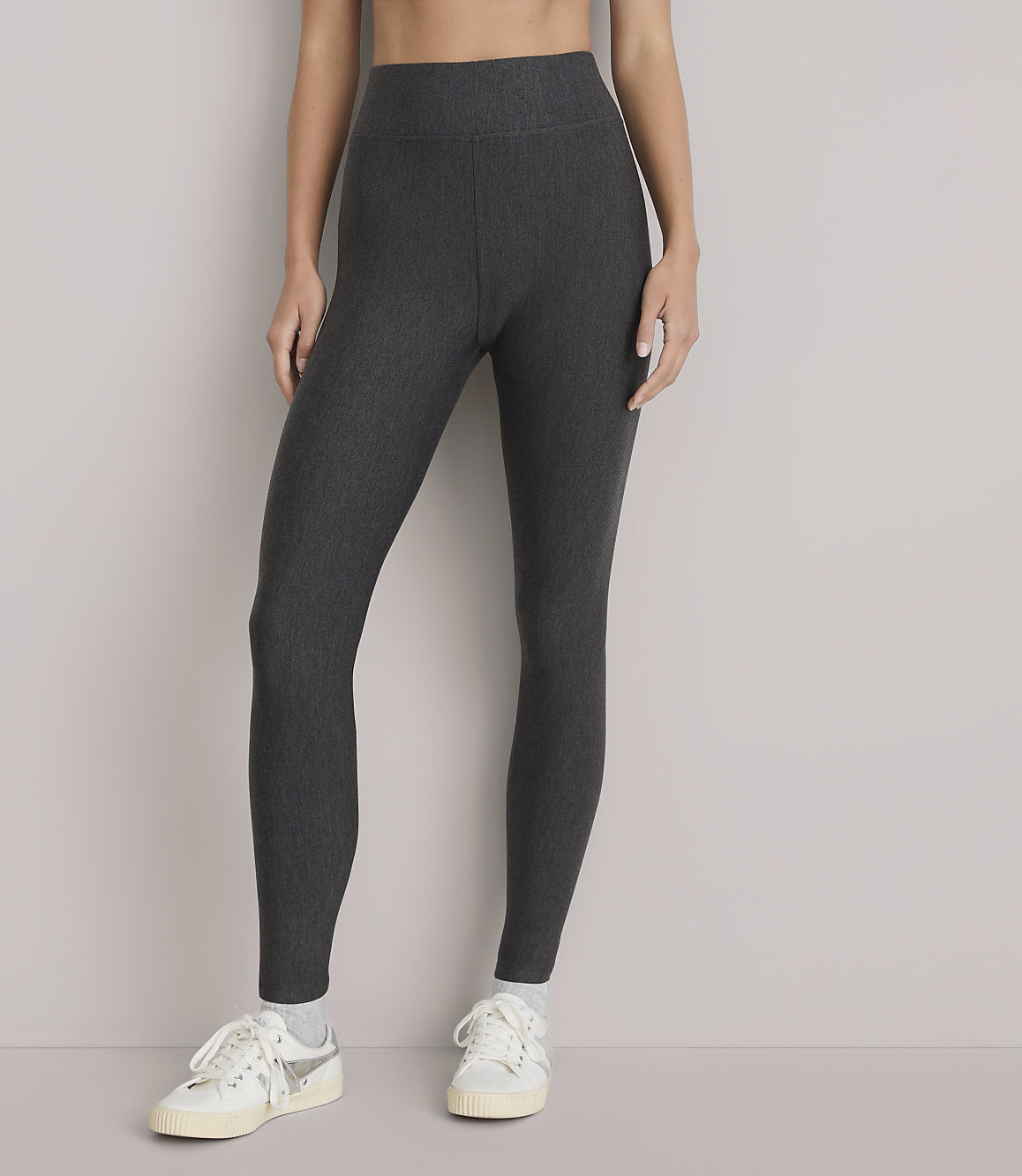 A person wearing dark grey ribbed leggings and white sneakers stands against a plain white background.