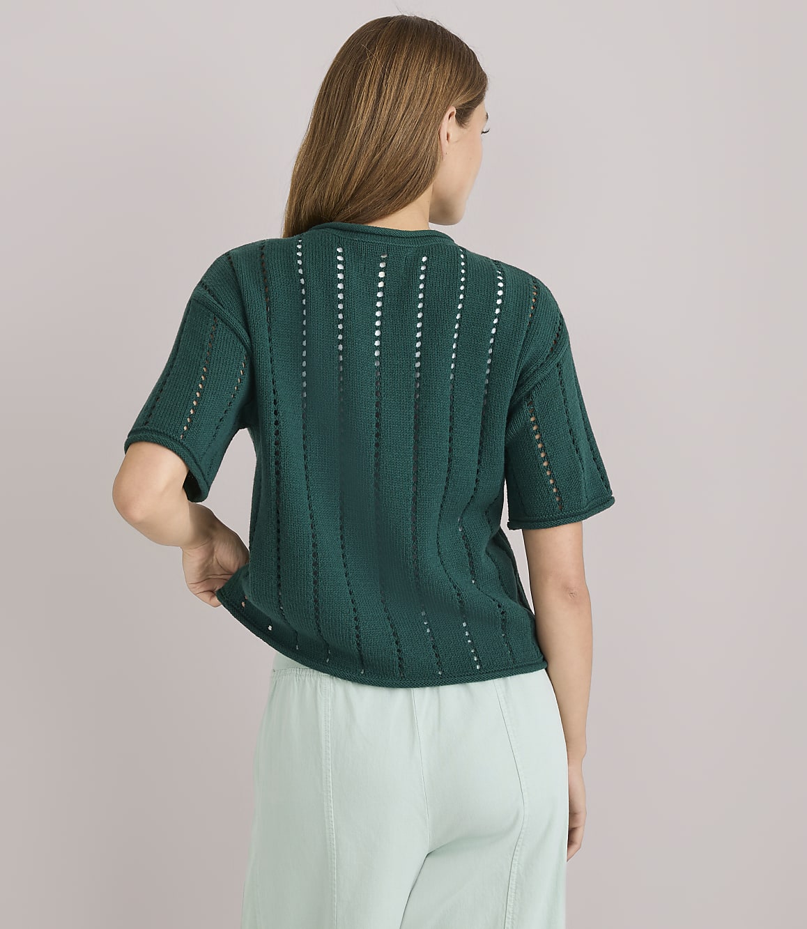 A woman in a green, short-sleeved, open-knit sweater and light green pants stands against a plain white background.