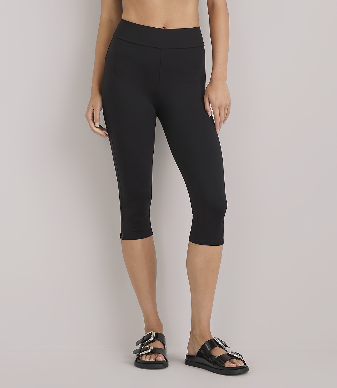 A person wearing black capri leggings and black sandals stands against a plain white background.
