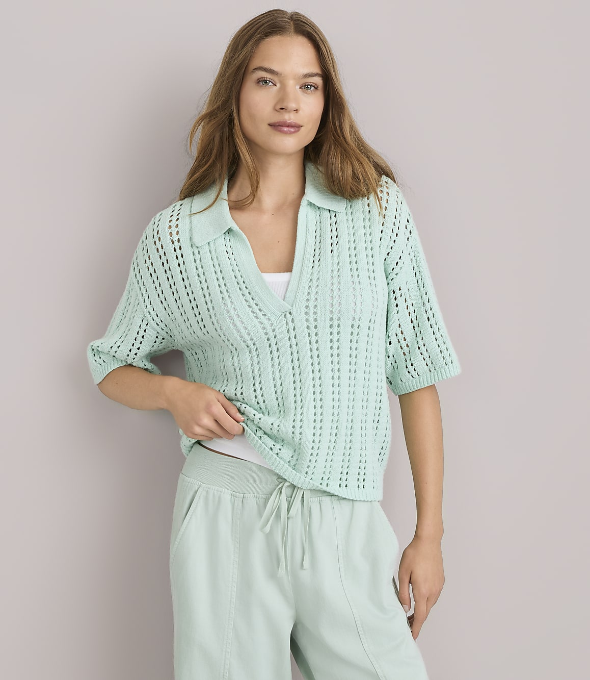 A woman in a light green collared knit shirt and matching sweatpants stands against a plain gray background.