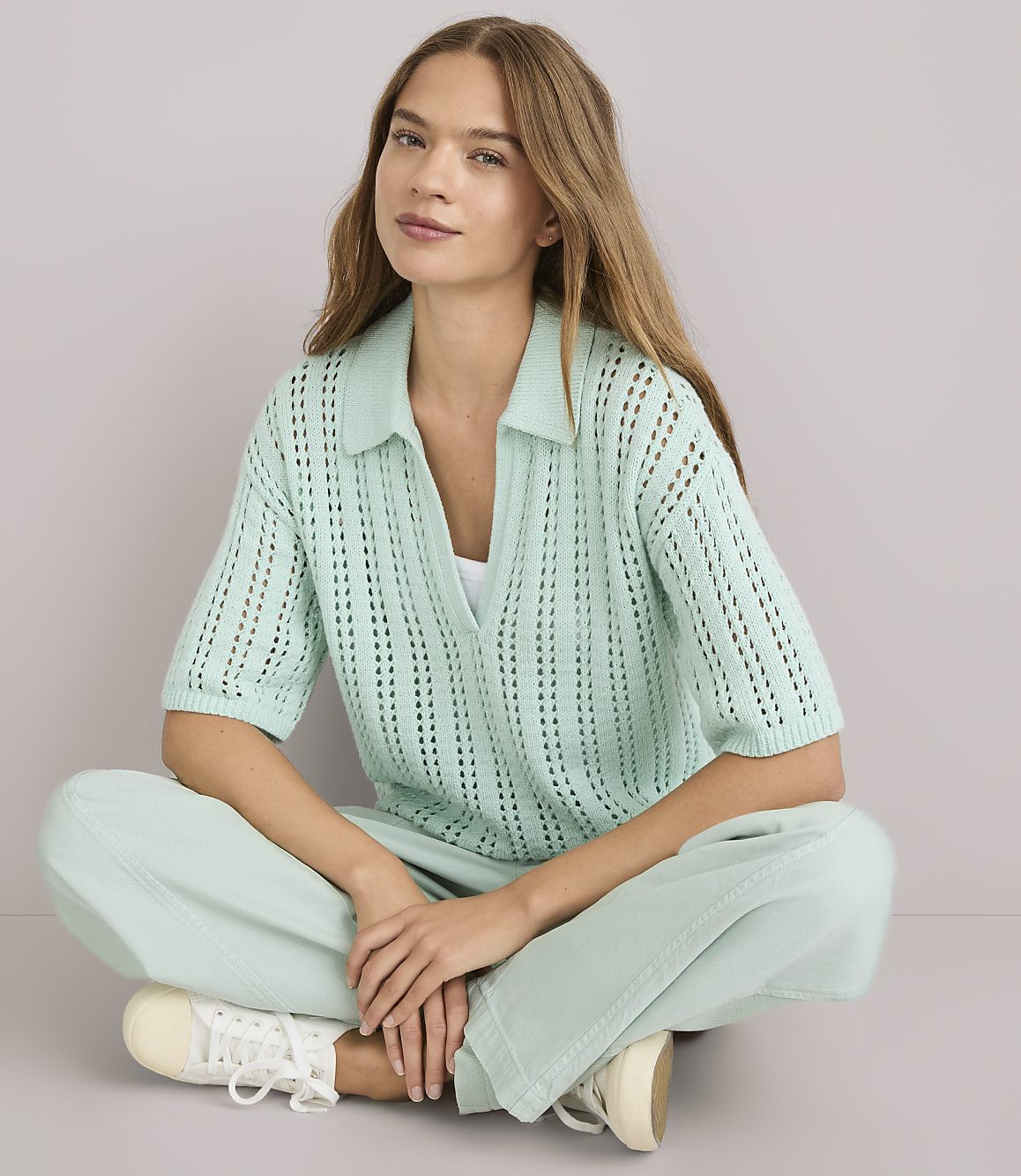 A young woman with long, light brown hair sits cross-legged in a light mint green collared knit shirt and matching wide-leg pants, against a plain white background.