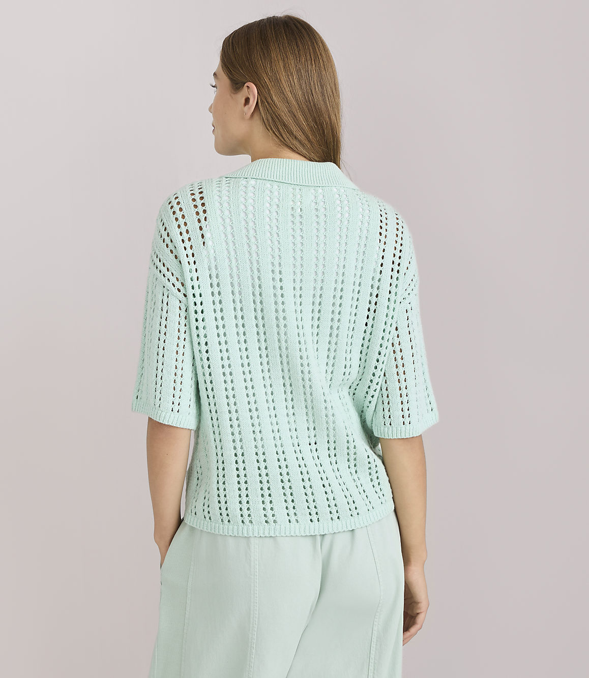 A woman in a light green collared knit shirt and matching pants stands against a plain white background.