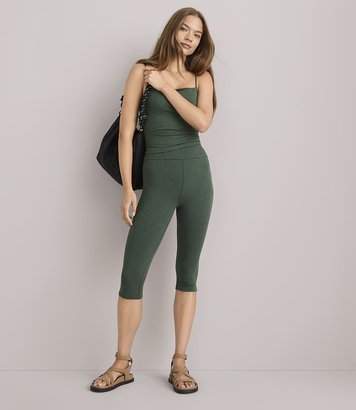 A woman in a green tank top and capri leggings stands with a black bag slung over her shoulder against a plain gray background.