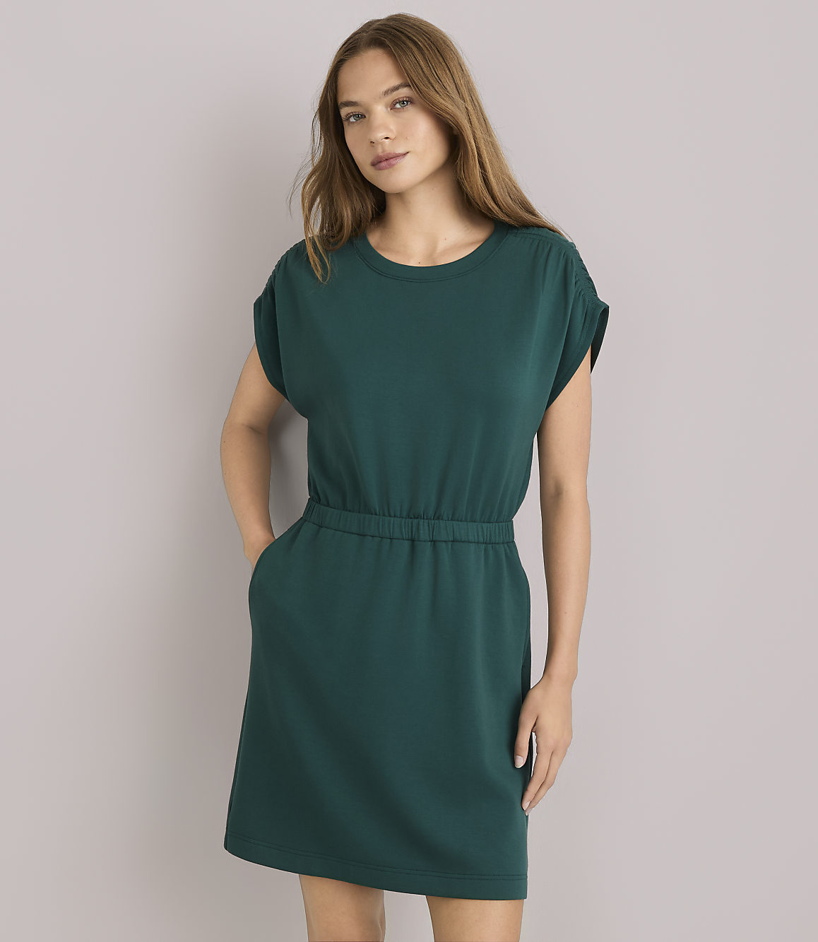 A woman in a green dress and sunglasses leans against a plain gray wall, with a light blue sweater tied around her waist.