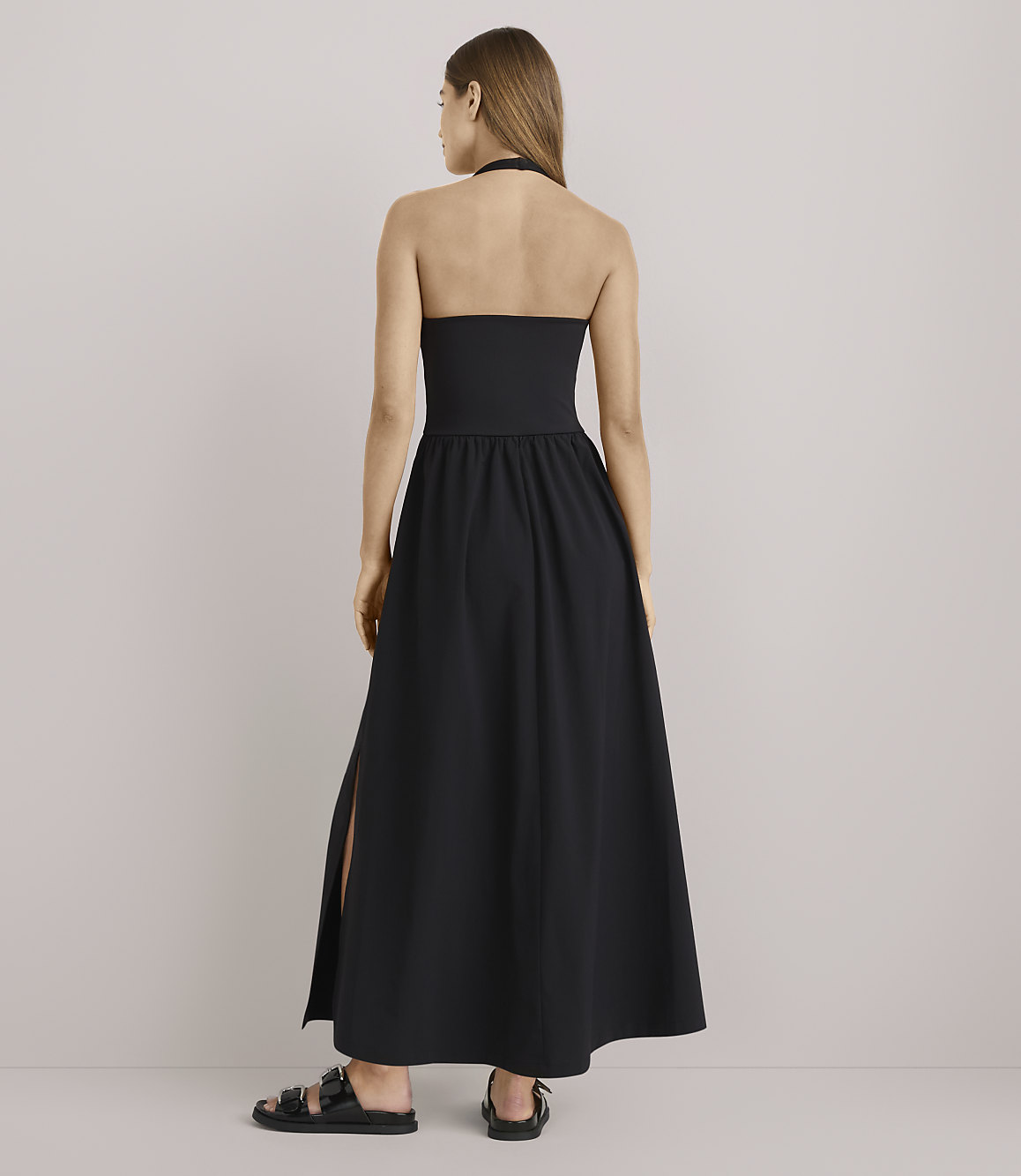 A woman in a black halter maxi dress with a slit stands against a plain white background.