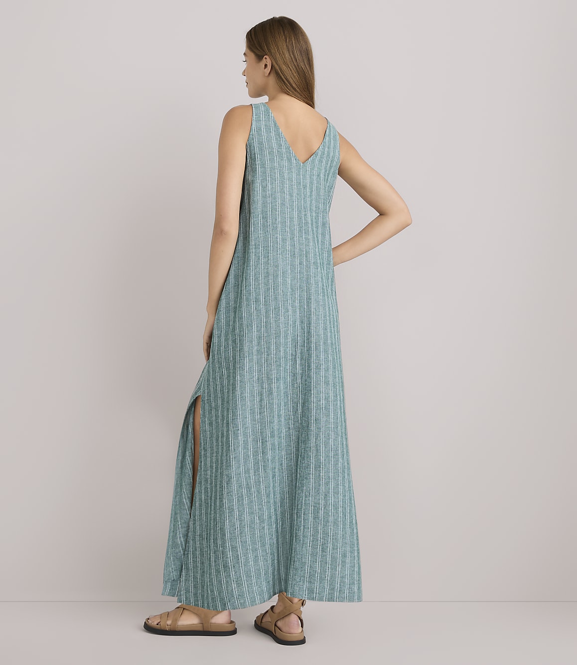 A woman in a teal striped maxi dress with side slits stands with her back to the camera against a plain white background.