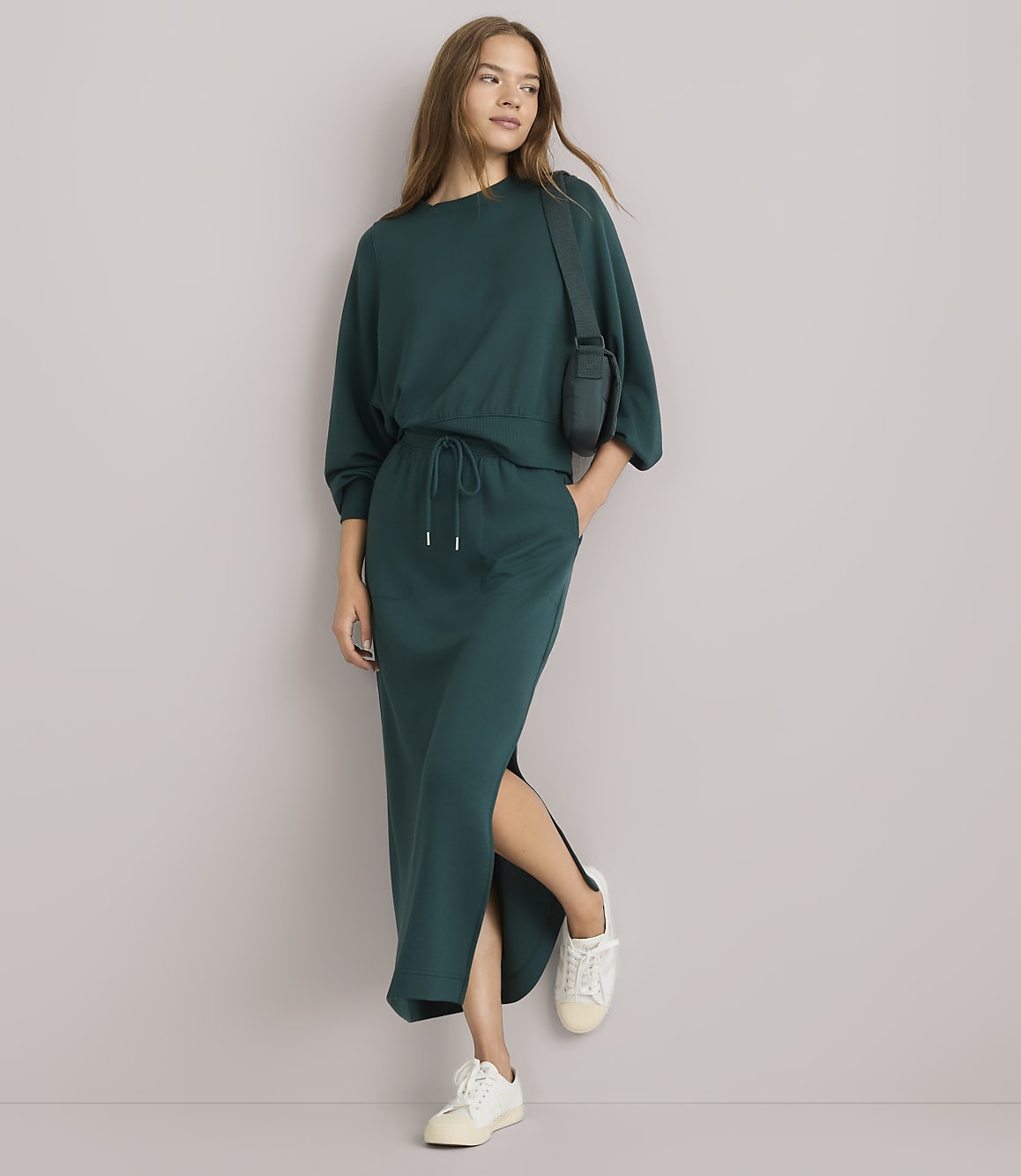 A woman in a teal sweatshirt and matching skirt with a side slit stands against a plain white background, wearing white sneakers and a small crossbody bag.