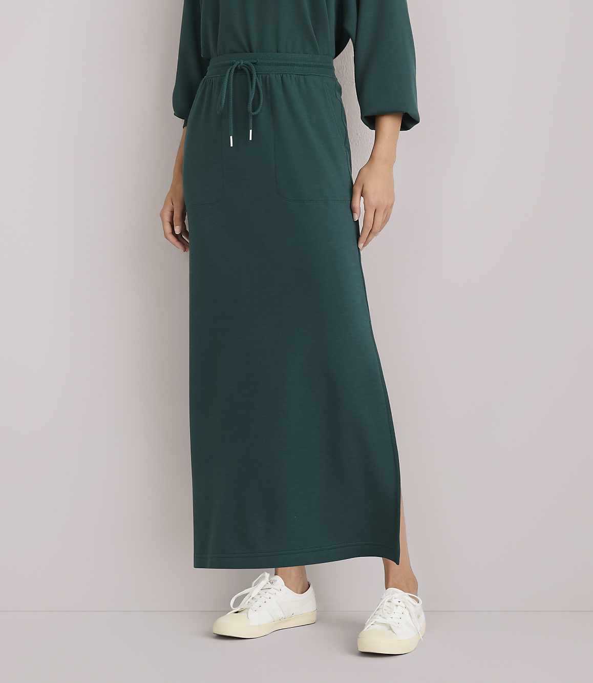 A person wearing a long, dark green dress with white sneakers stands against a plain white background.
