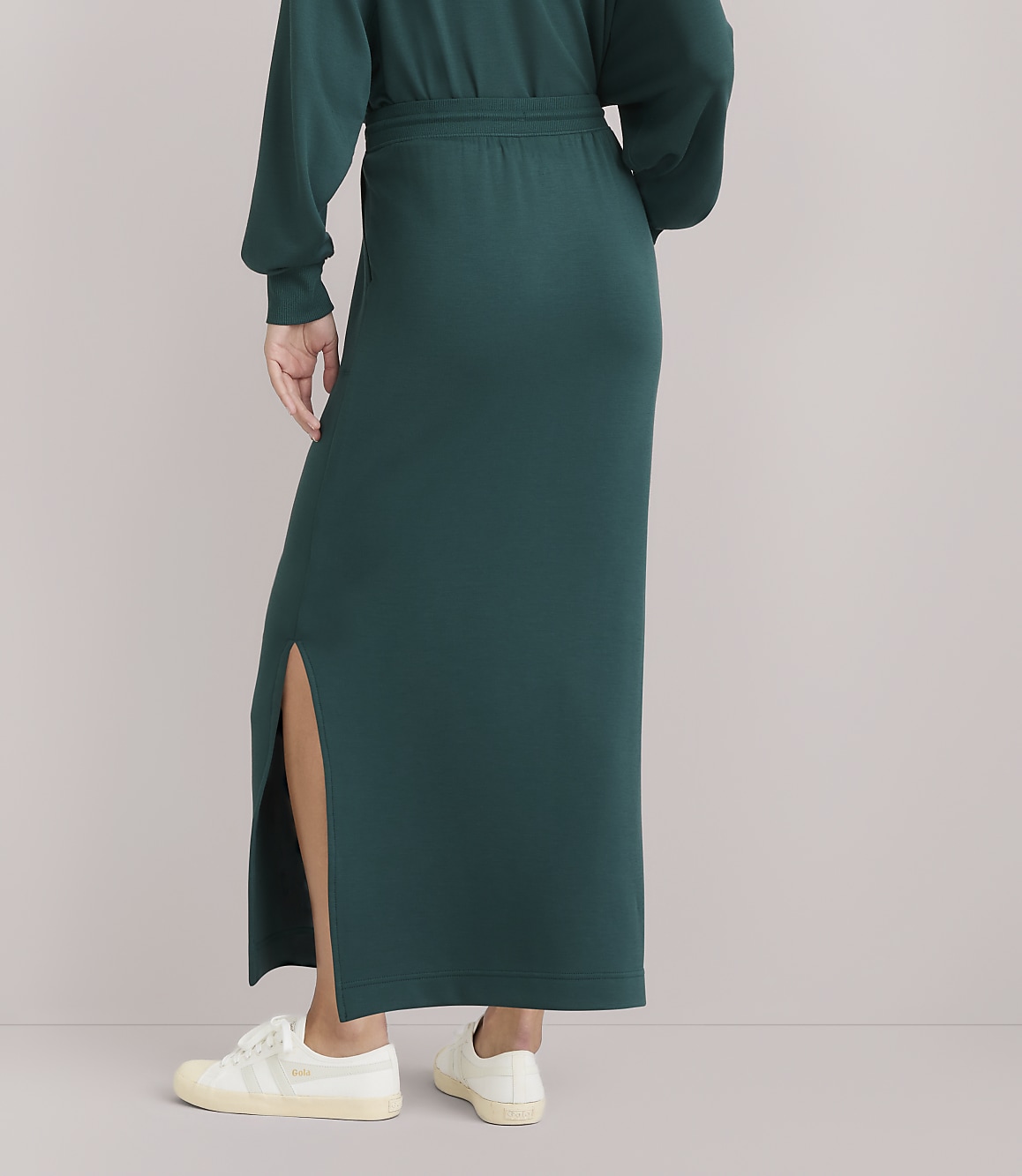A person in a long, dark green skirt with a high slit and white sneakers stands against a plain white background.