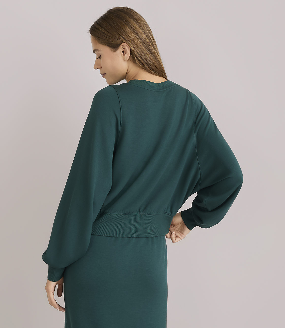 A woman in a teal sweatshirt with voluminous sleeves stands against a plain, light gray background.