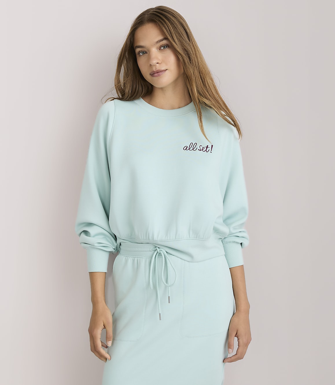 A young woman with light brown hair, wearing a light blue sweatshirt with "all set!" embroidered on it, poses with her arm behind her head against a plain white background.