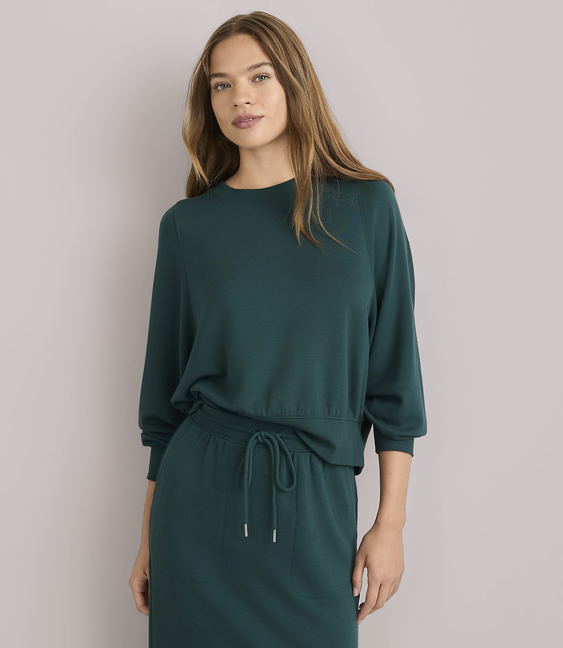 A young woman with long brown hair wears a dark green sweater and skirt, sitting against a plain white background.