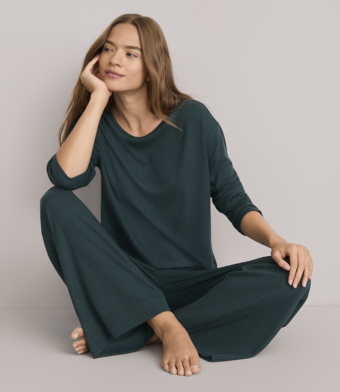 A woman in a dark green ribbed pajama set sits cross-legged on a plain white floor, with a soft, neutral background.