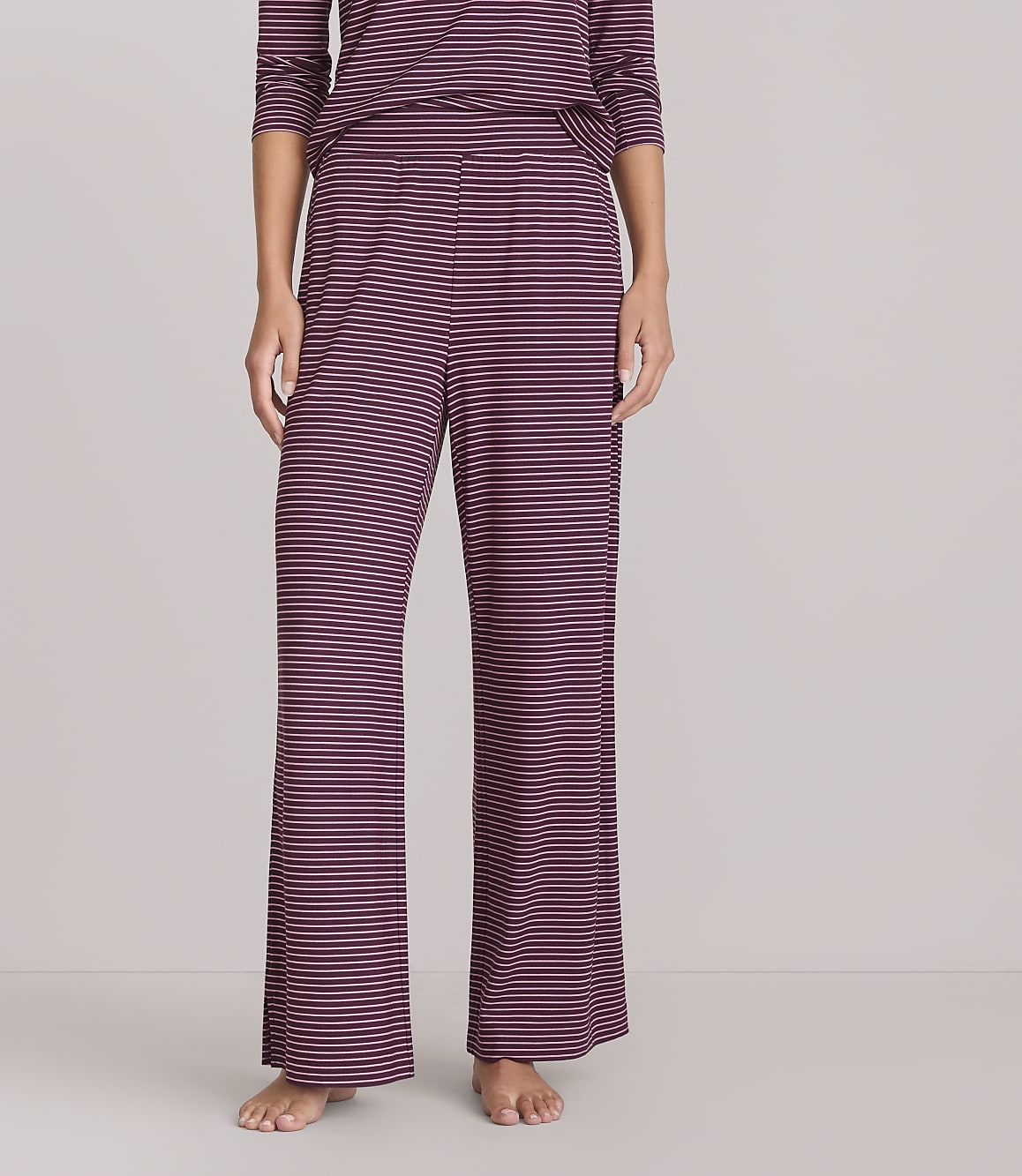 A person stands wearing burgundy and navy striped pajama pants and a matching top against a plain white background.