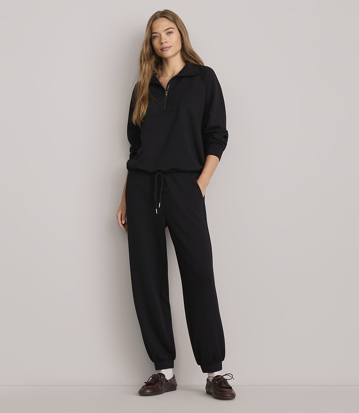 A woman in a black tracksuit stands against a plain white background.