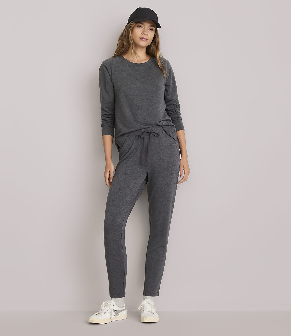 A woman in a dark gray loungewear set and a black baseball cap stands against a plain white background.