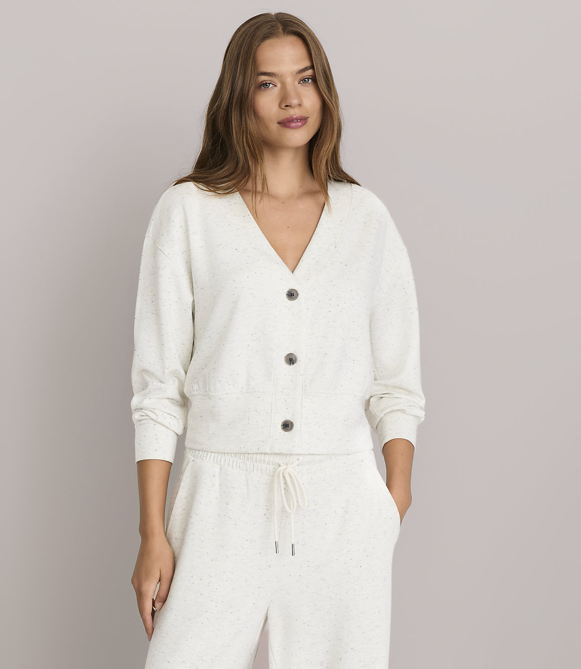 A woman in a cream-colored cardigan and matching pants stands against a plain gray background.
