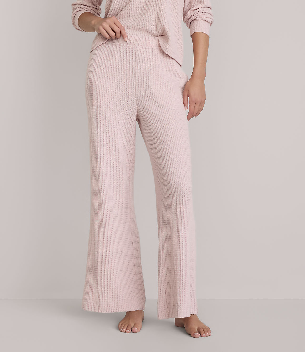 A person stands barefoot in pink waffle-knit pajama pants and a matching top against a plain white background.