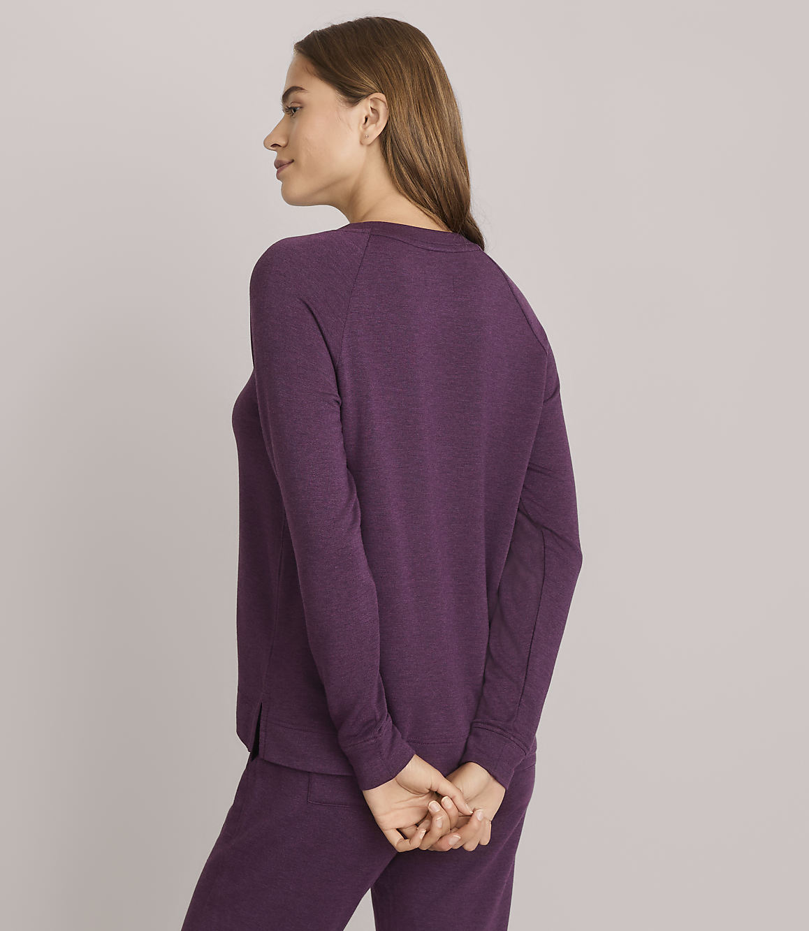 A woman in a purple sweatshirt and pants stands with her hands clasped behind her back against a plain gray background.