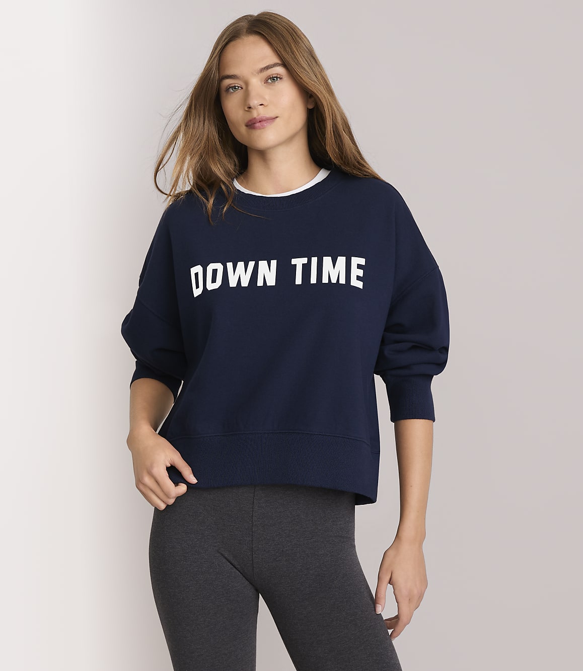 A young woman in a navy sweatshirt with "DOWN TIME" printed on it and dark leggings stands against a plain white background.