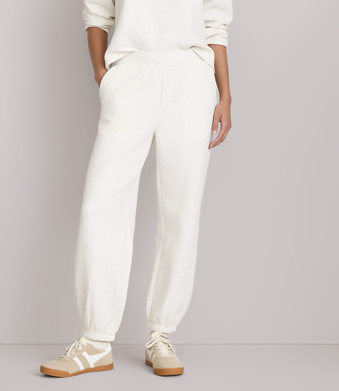 A person wearing cream-colored waffle-knit sweatpants and sneakers stands against a plain, light gray background.