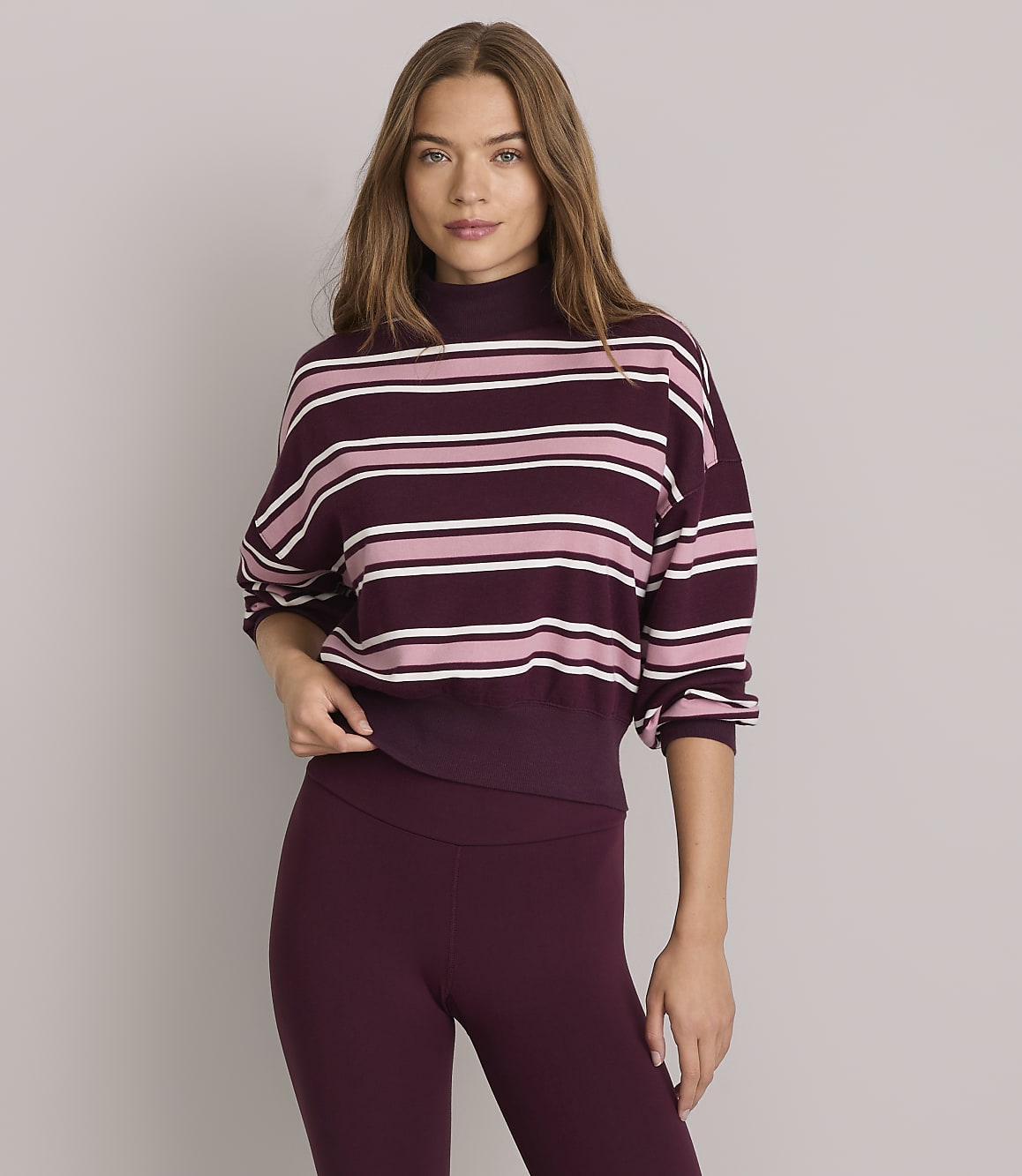 A woman in a striped mock-neck sweatshirt and burgundy leggings stands against a plain gray background.