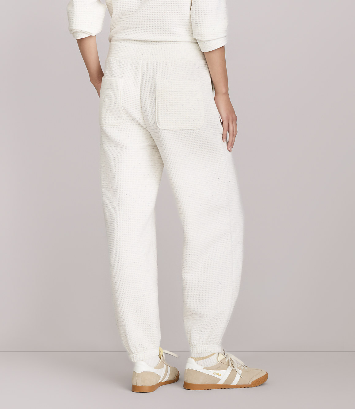 A person wearing cream-colored waffle-knit sweatpants and sneakers stands against a plain white background.